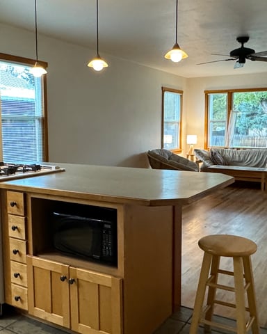 Open-plan kitchen and living room with wood cabinetry, island, gas stove, and seating area with large windows overlooking trees.