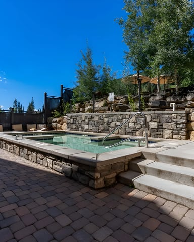Hot spring pool with stone surround, lounge chairs, and mountain landscape under clear blue sky
