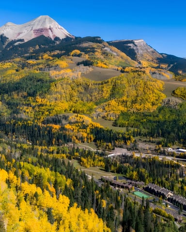 Mountain valley with snow-capped peak, golden aspen trees, evergreen forests, and resort buildings nestled in the landscape under clear blue sky