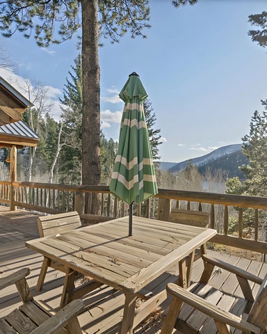 Log cabin deck with wooden furniture, green striped umbrella, and mountain lake view surrounded by pine trees