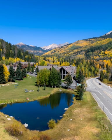 Mountain resort with brown buildings, small pond, highway, and snow-capped peak in autumn landscape with golden aspen trees