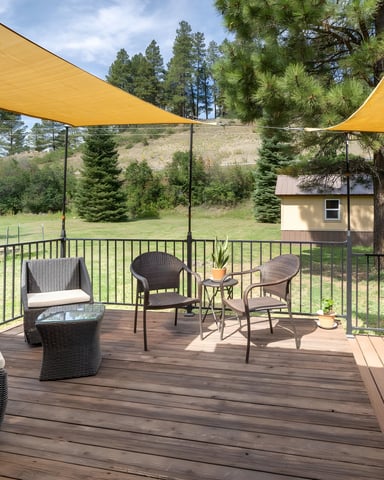 Spacious deck with yellow shade sails, wicker seating, grill, and views of forested hillside and small outbuilding.