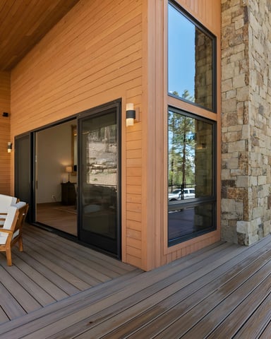 Modern wood and stone cabin exterior with wraparound deck, lounge chairs, large windows, and mountain forest views