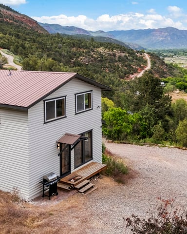 Modern white house with red metal roof on hillside overlooking forested valley and distant mountains