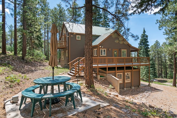 Modern cabin with metal roof and wraparound deck nestled among tall pine trees, picnic table in foreground