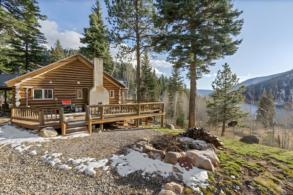 Log cabin with stone chimney and wraparound deck surrounded by tall pine trees and mountains in background
